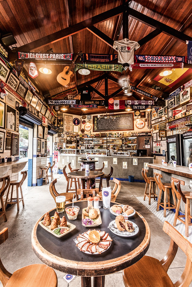 Salão do Bar do Luiz Fernandes com mesa posta com petiscos à frente em destaque, bancada ao fundo e banquetas em ambos os lados.